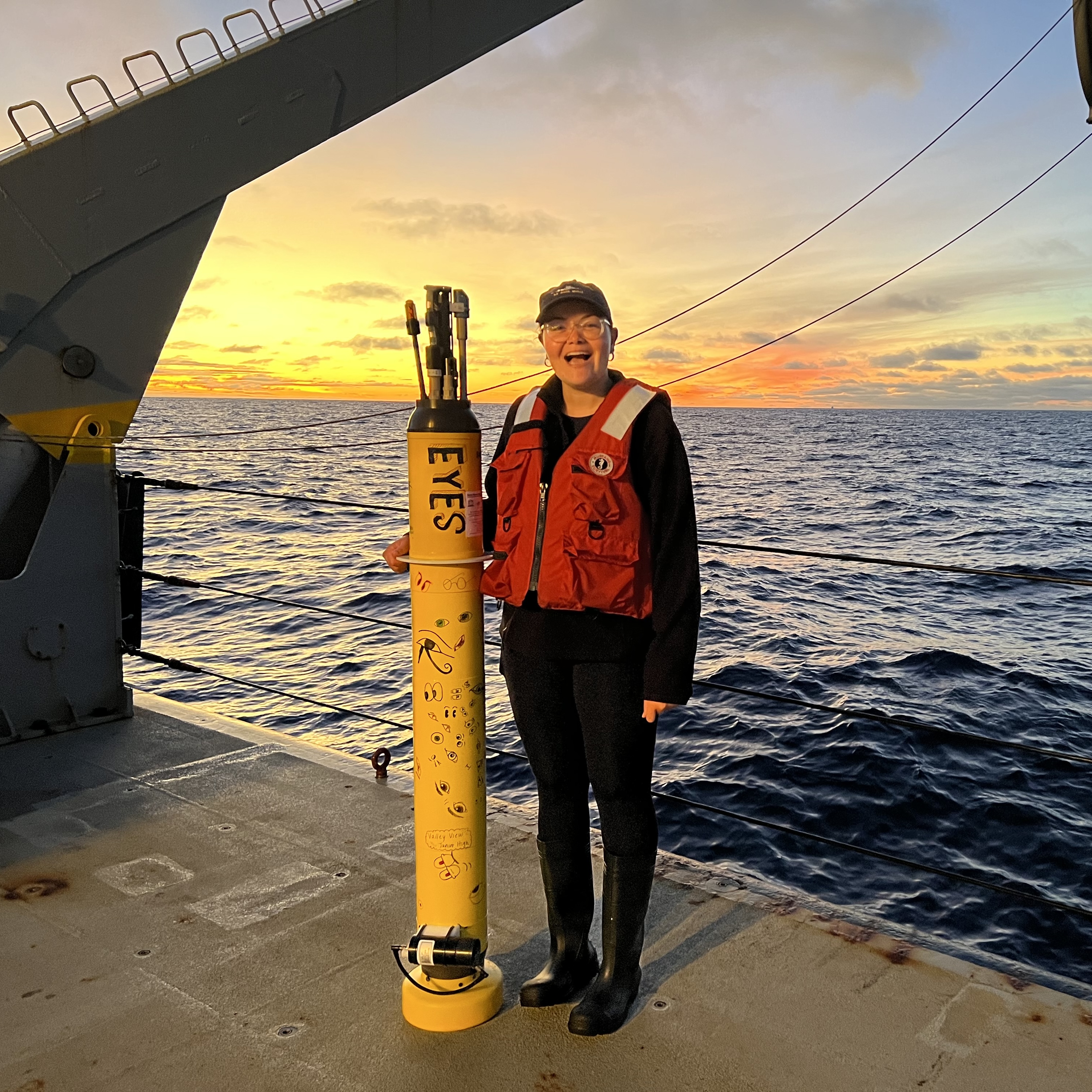 Lauren wearing orange life jacket, standing on ship deck next to yellow float at sea at sunset