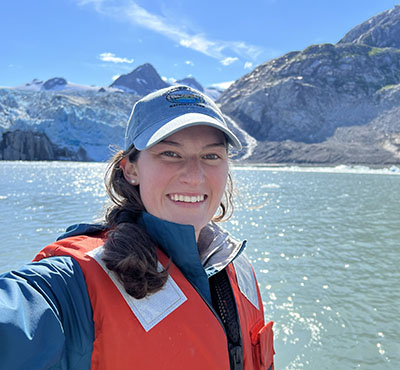 Mary Margaret in hat and orange life vest in front of water and icy mountains