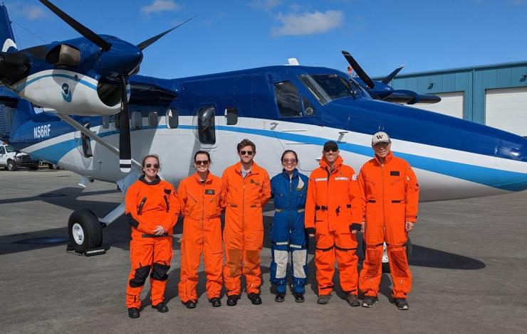 The aerial flight team of six pose in front of a blue and white NOAA Twin Otter aircraft wearing flight suits in both orange and blue. 