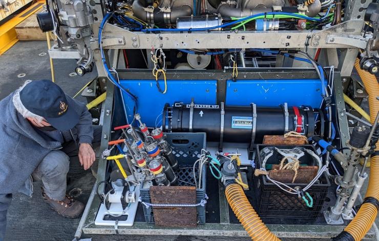 Dave squatting to the left of a remotely operated vehicle with a blue hat and mask on. Variety of tubes, bottles and sensors are shown as part of the ROV