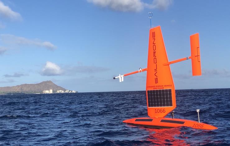 Red saildrone floats on ocean waves under blue skies with Diamond Head, Hawaii and buildings in distance