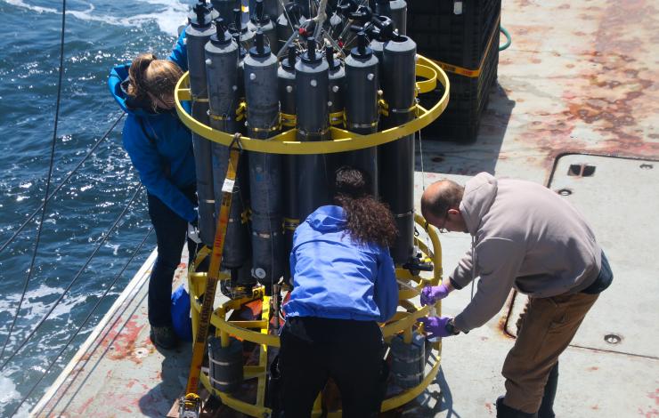 People stand around a circular CTD instrument collecting water samples on a ship out in the Pacific Ocean