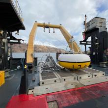 A yellow and white painted circular donut shaped mooring upside down on the back of a ship with the sea and landscape in the background