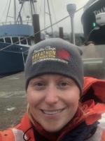 Angie is orange safety jacket with grey beanie smiling in front of a dock with a blue research ship